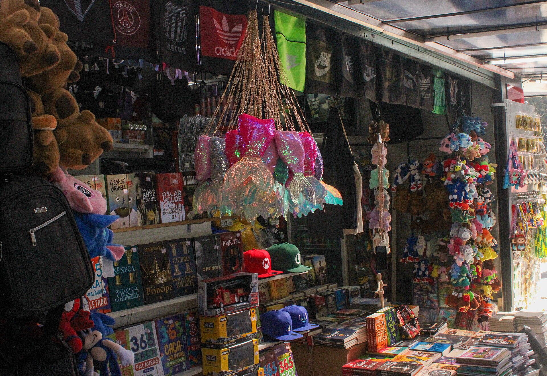 Interior de uma banca de jornal, com livros, pelúcias, bonés e brinquedos nas prateleiras